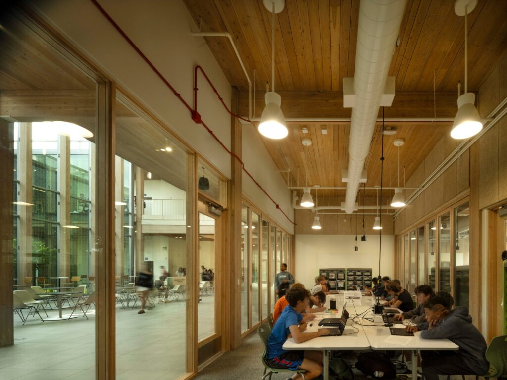 Students working with laptops on long tables inside a brightly lit STEM classroom with wood ceilings and glass partitions.