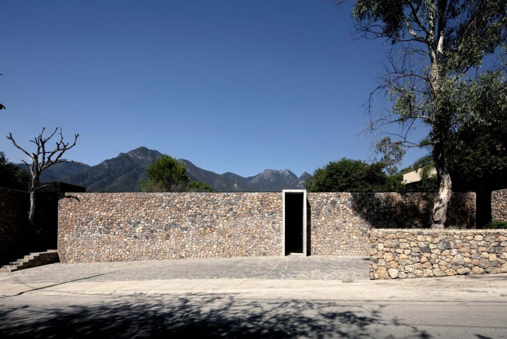 A massive local stone wall with a minimalist vertical entrance under a clear blue sky with mountains in the background.