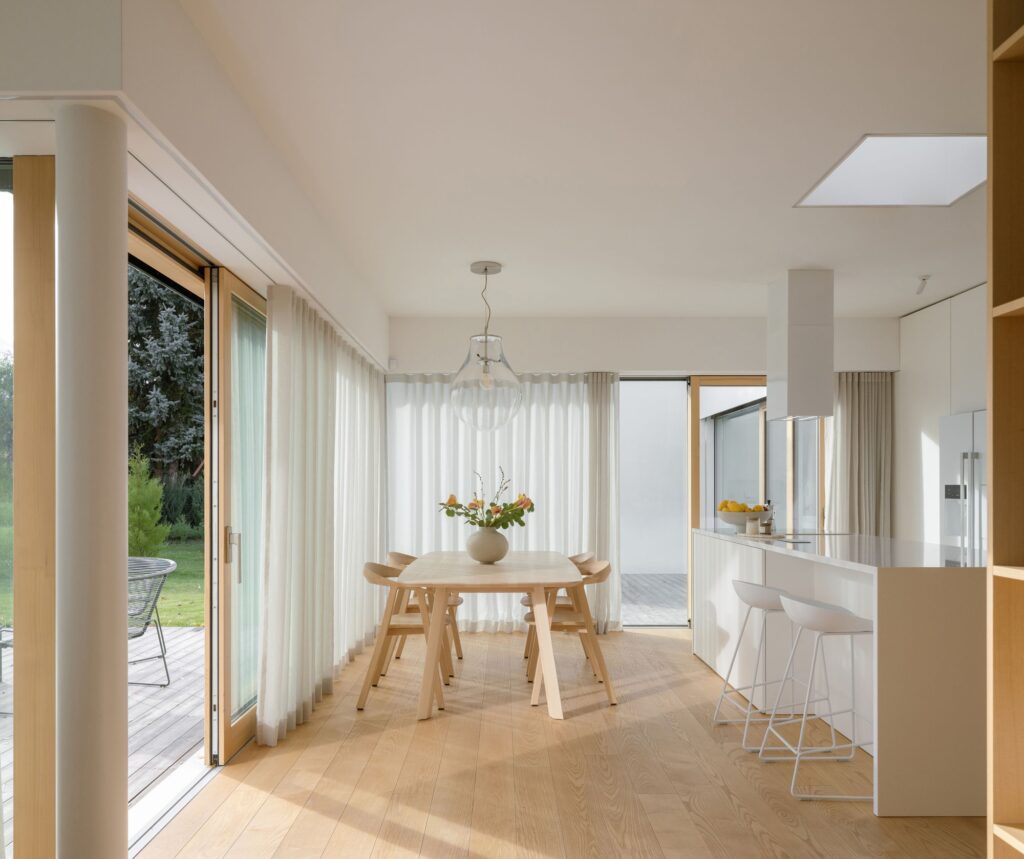 Bright interior dining area and minimalist white kitchen of Sestajovice House with natural wood flooring and soft daylight.