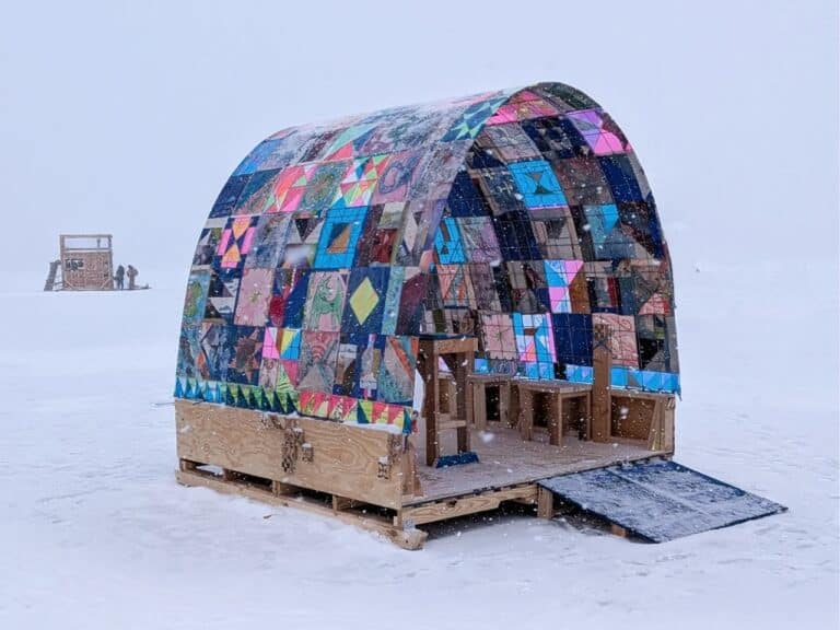 A quilted arched structure known as an Art Shanty on a frozen lake during a snowfall, featuring colorful patchwork patterns and a wooden base with a ramp.