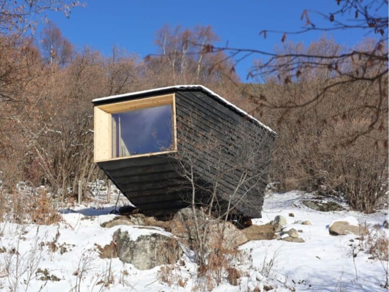 Exterior view of the Forestone cabin in the Pyrenees mountains during winter, featuring black Yakisugi wood cladding and a large panoramic window.