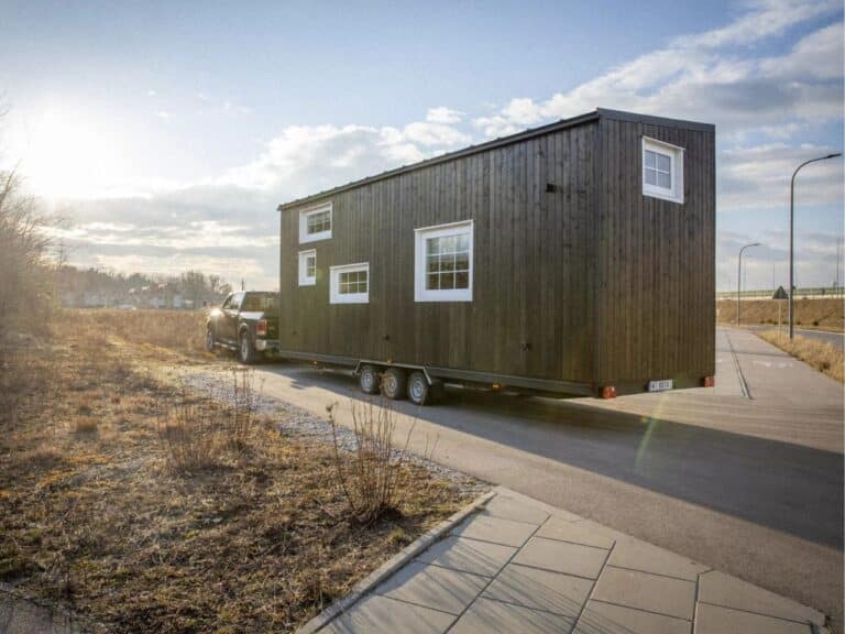 Exterior view of a modern dark wood tiny house on a triple-axle trailer being towed by a pickup truck on a highway.