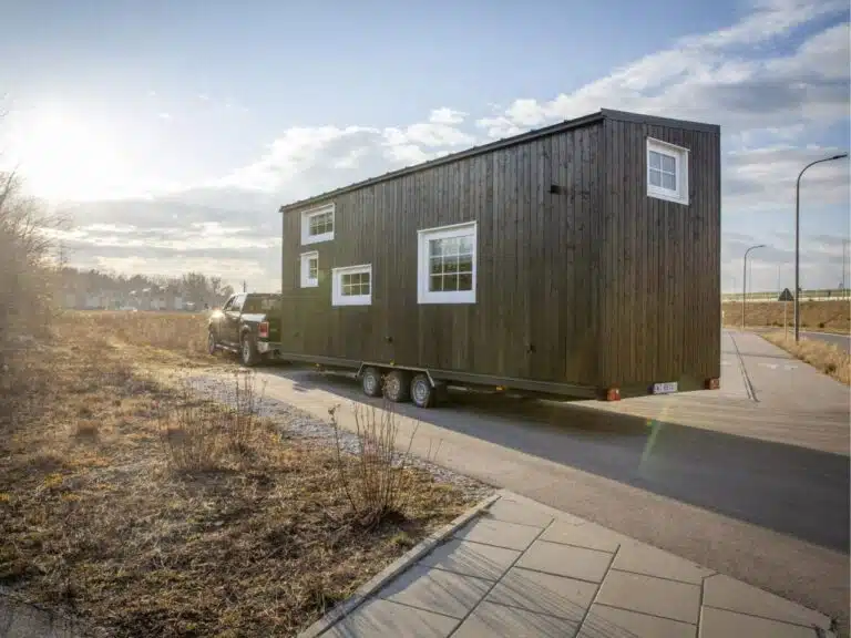 Exterior view of a modern dark wood tiny house on a triple-axle trailer being towed by a pickup truck on a highway.