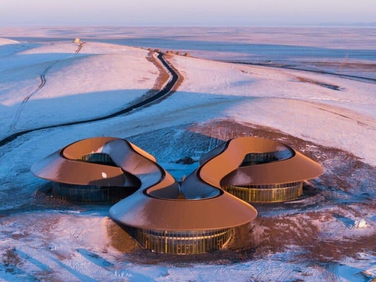 Aerial view of the Volcano Visitor Center in China during sunset, showing three circular volumes connected by a continuous curved roof on a snowy landscape.