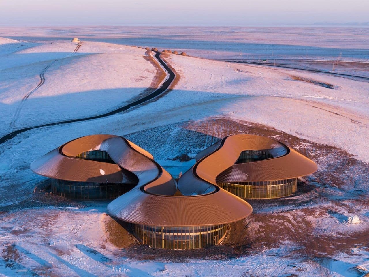 Aerial view of the Volcano Visitor Center in China during sunset, showing three circular volumes connected by a continuous curved roof on a snowy landscape.