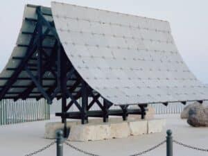 A temporary architectural pavilion with a pitched roof made of porous grey Etna lava stone tiles supported by a black charred wood frame on limestone blocks.