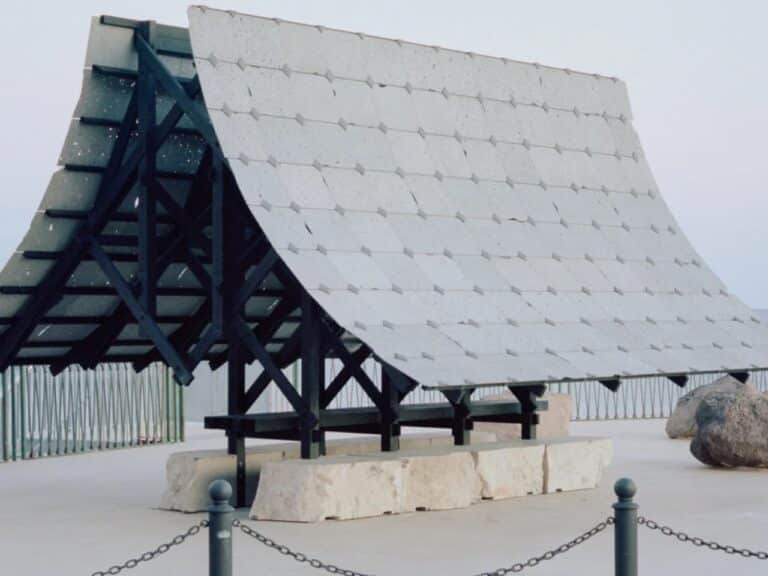 A temporary architectural pavilion with a pitched roof made of porous grey Etna lava stone tiles supported by a black charred wood frame on limestone blocks.