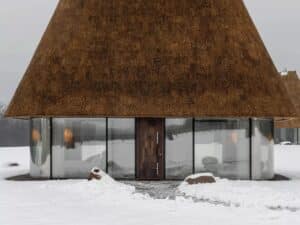 Front view of a modern Ukrainian Mazanka house with floor-to-ceiling glass walls and a thick sculptural thatched roof in a snowy landscape.