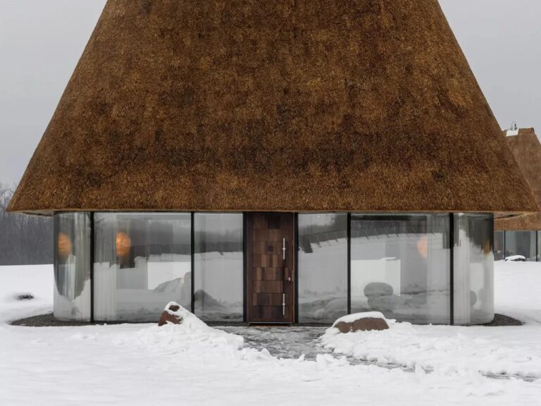 Front view of a modern Ukrainian Mazanka house with floor-to-ceiling glass walls and a thick sculptural thatched roof in a snowy landscape.