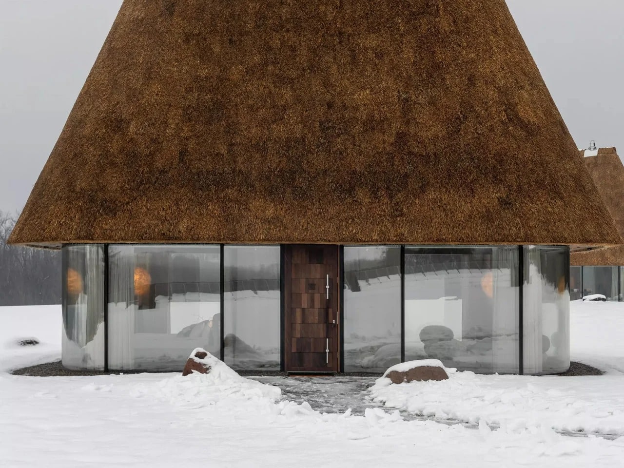Front view of a modern Ukrainian Mazanka house with floor-to-ceiling glass walls and a thick sculptural thatched roof in a snowy landscape.