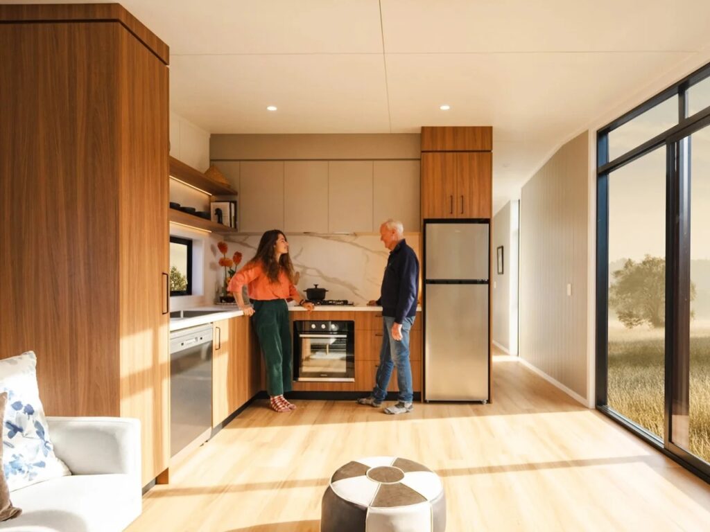A spacious open-plan interior of a tiny house with wood-textured cabinets, a marble backsplash, and large glass doors.