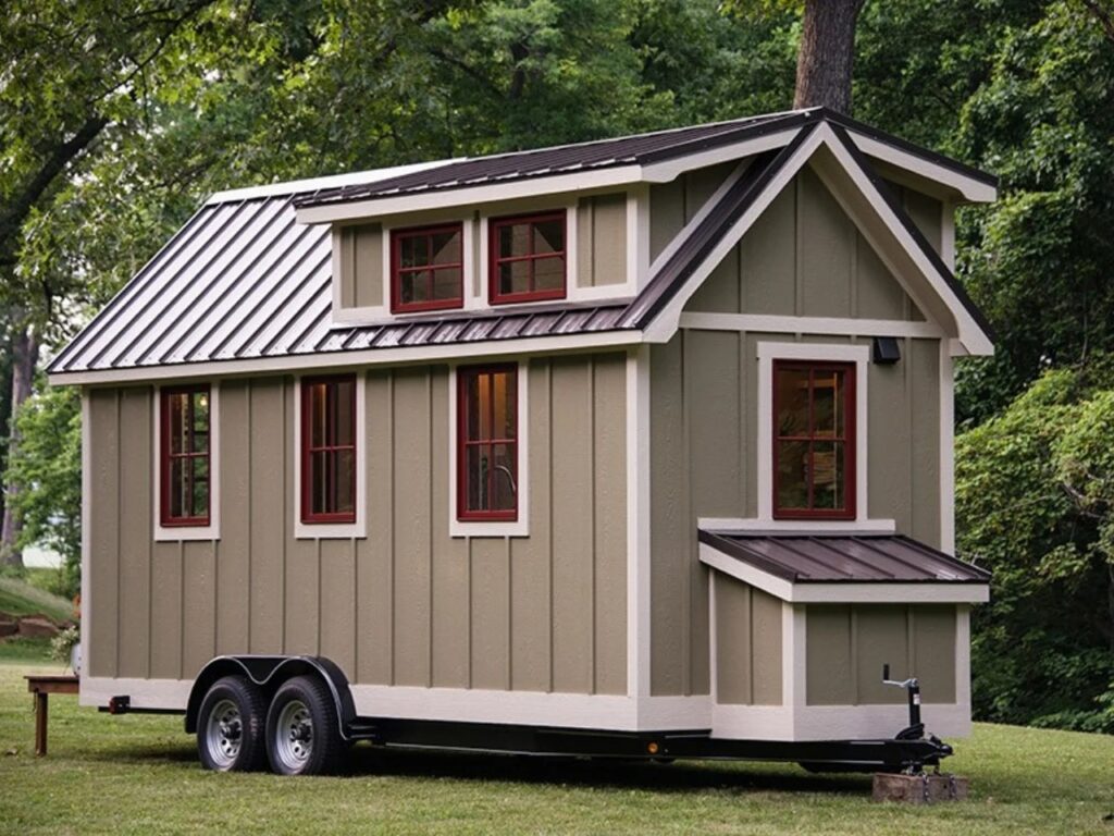 Side and rear exterior view of the Ynez tiny house showing multiple windows with red frames, green-toned siding, and the trailer chassis.