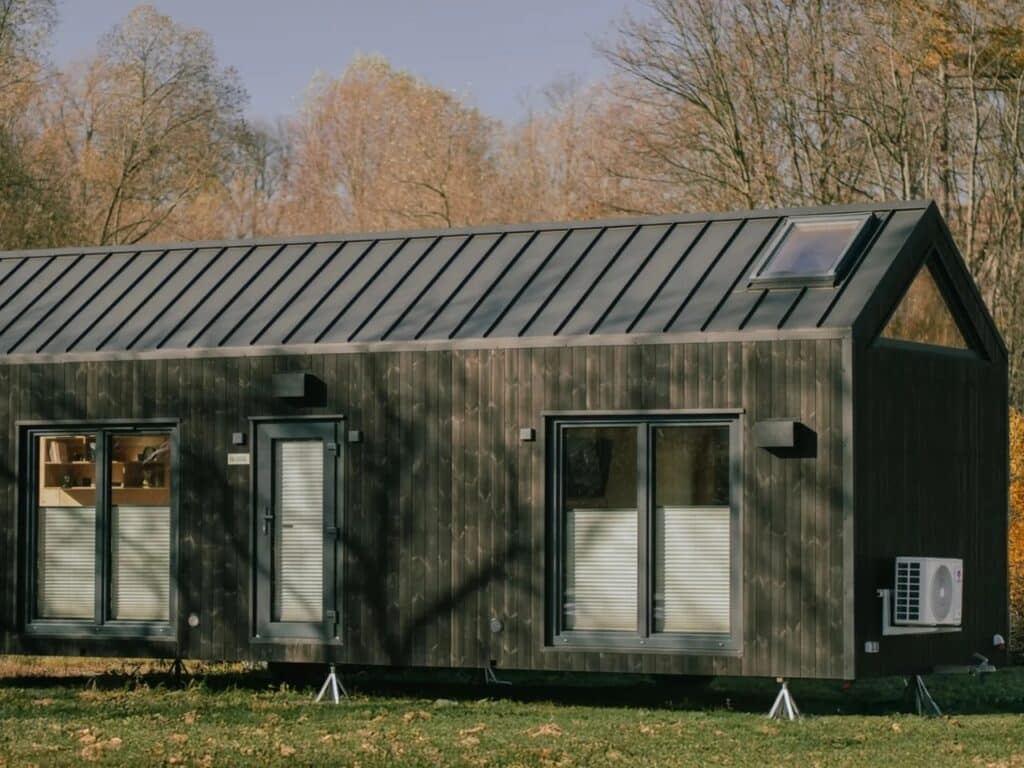 Side view of Zenith tiny house on a grassy field, showing double glass doors, windows with blinds, and an external HVAC unit.
