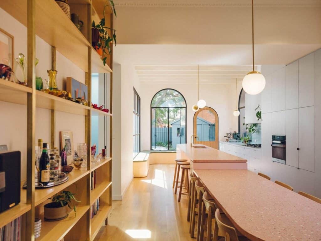 Wide view of the open-plan interior of the Adelaide cottage, featuring a long pink kitchen island, timber shelving, and arched garden doors.