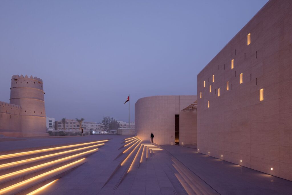 Al Ain Museum at dusk featuring illuminated linear steps, modern stone buildings, and the historic fort under a purple sky.