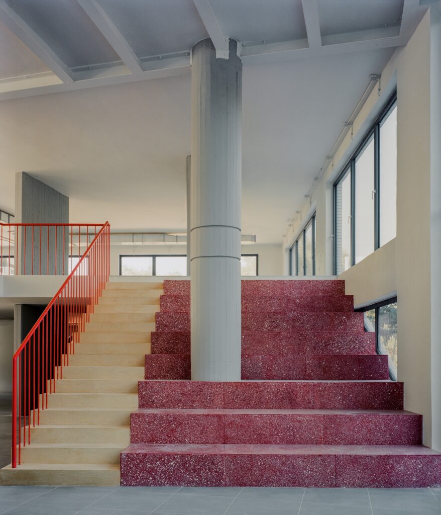 A dual-material staircase featuring light wood steps on the left and red terrazzo steps on the right, divided by a minimalist red railing.