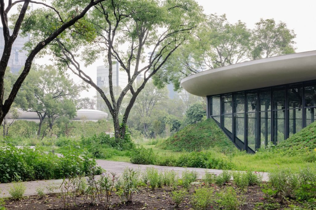 A glass-walled pavilion surrounded by lush green mounds and native vegetation with a walking path in the foreground.