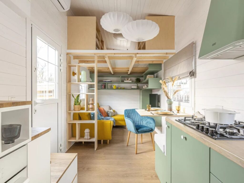 Interior of a tiny house showing a multifunctional living area with a yellow sofa, a blue chair, and a loft bedroom above.
