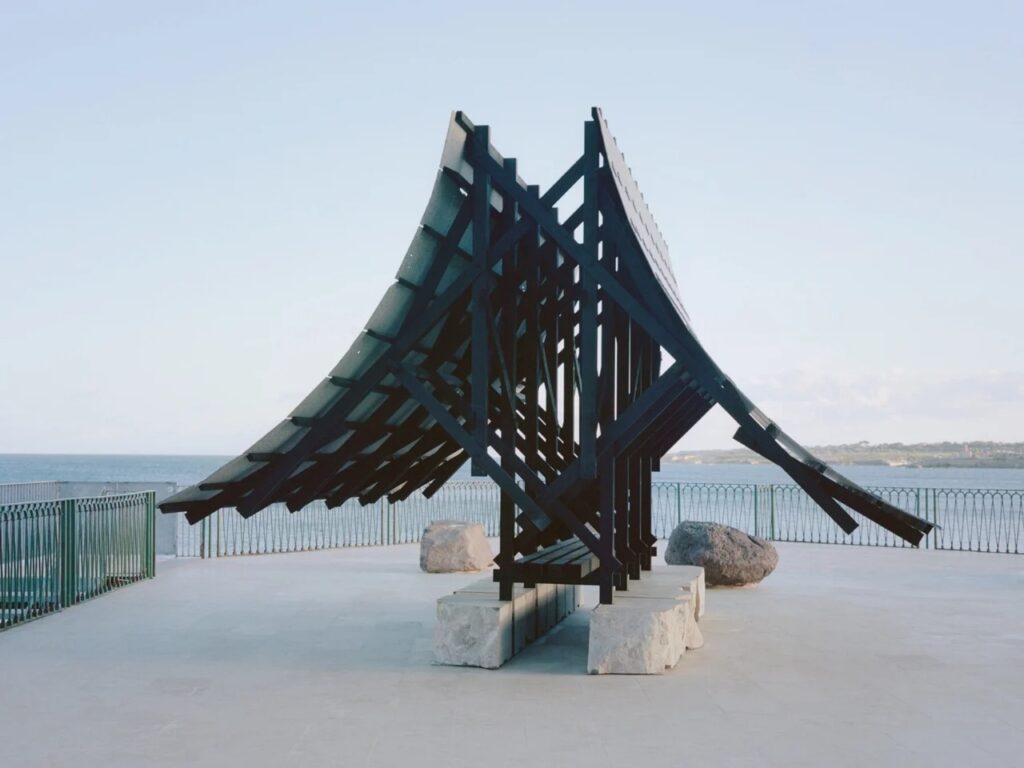 Front view of the Asympta installation showing two converging curved roof planes forming a bridge-like silhouette against a coastal background.