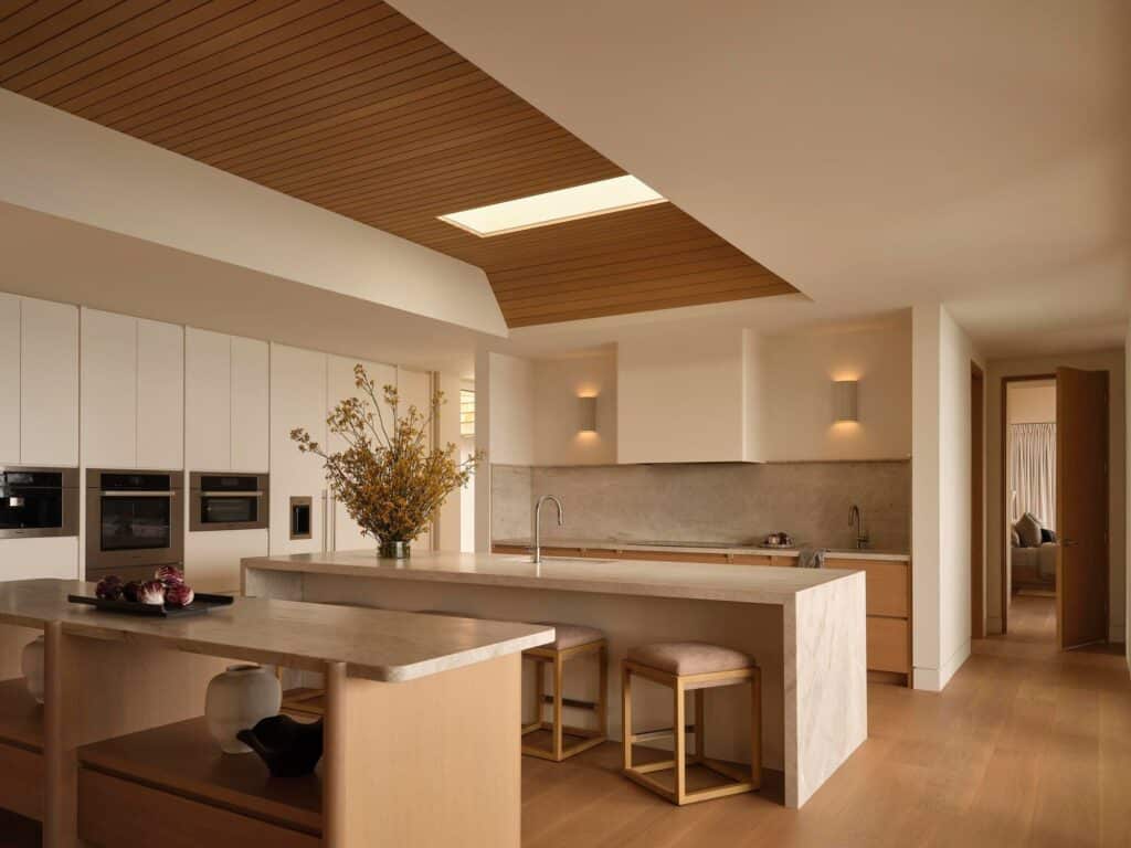 Minimalist kitchen design featuring a large stone island, light wood cabinetry, and integrated high-end appliances under a skylight.