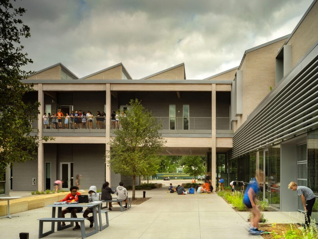 Students gathered in an outdoor courtyard between building wings, with a second-story walkway and saw-tooth rooflines visible.