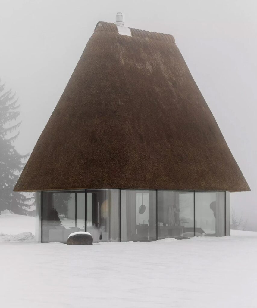 Side view of a small cabin with a tall, mushroom-like reed roof and transparent glass base during a foggy winter day.