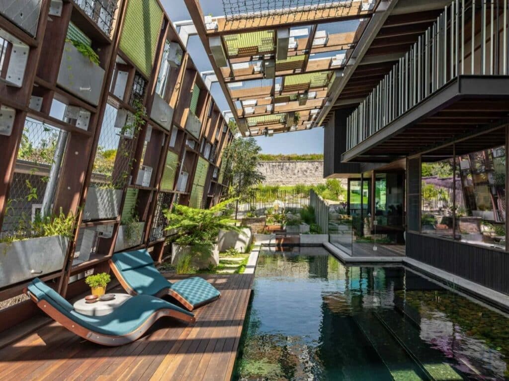 A long natural swimming pool integrated into the heart of Holocene House with a wooden deck, lounge chairs, and a vertical garden wall under a geometric timber canopy.