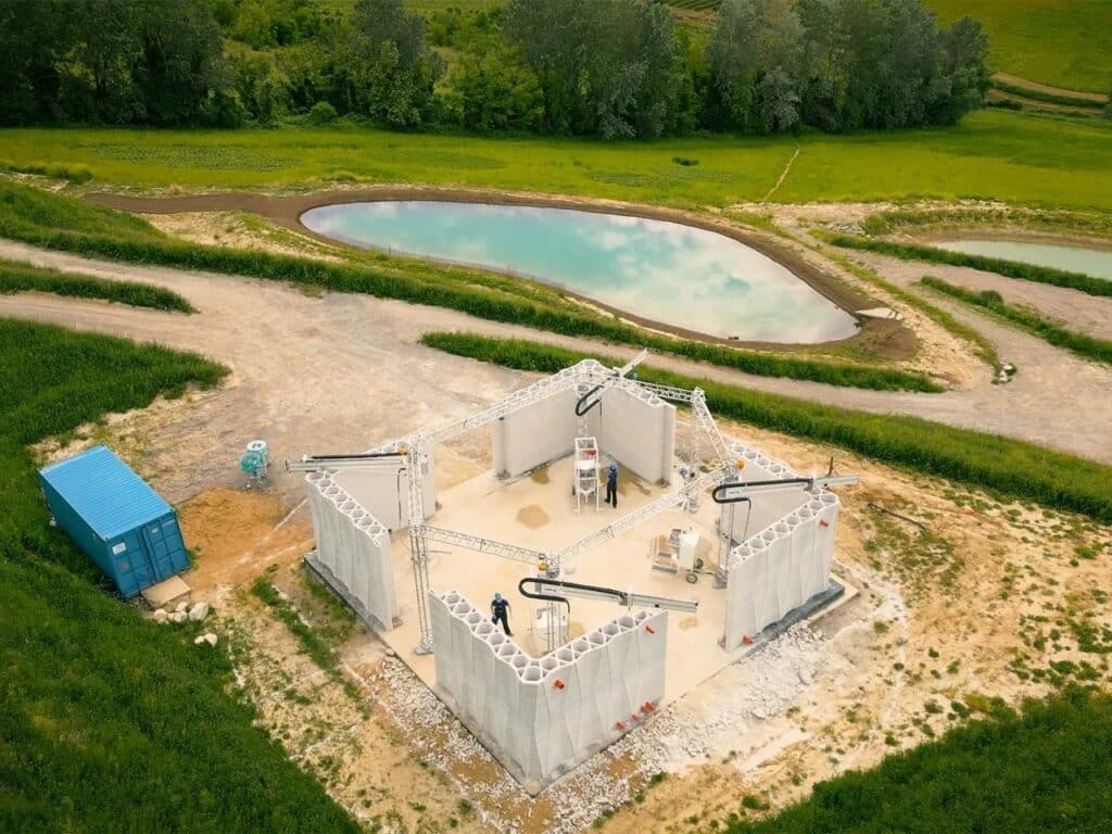 Wide aerial shot of the 3D-printed house under construction next to a natural pond in a green landscape in Italy.