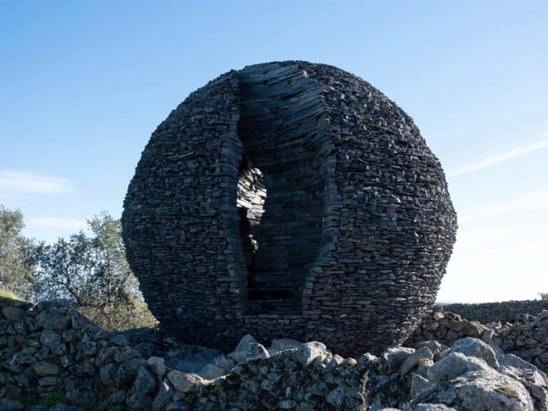A large spherical structure made of stacked dark schist stone with a narrow vertical opening, set against a clear blue sky.