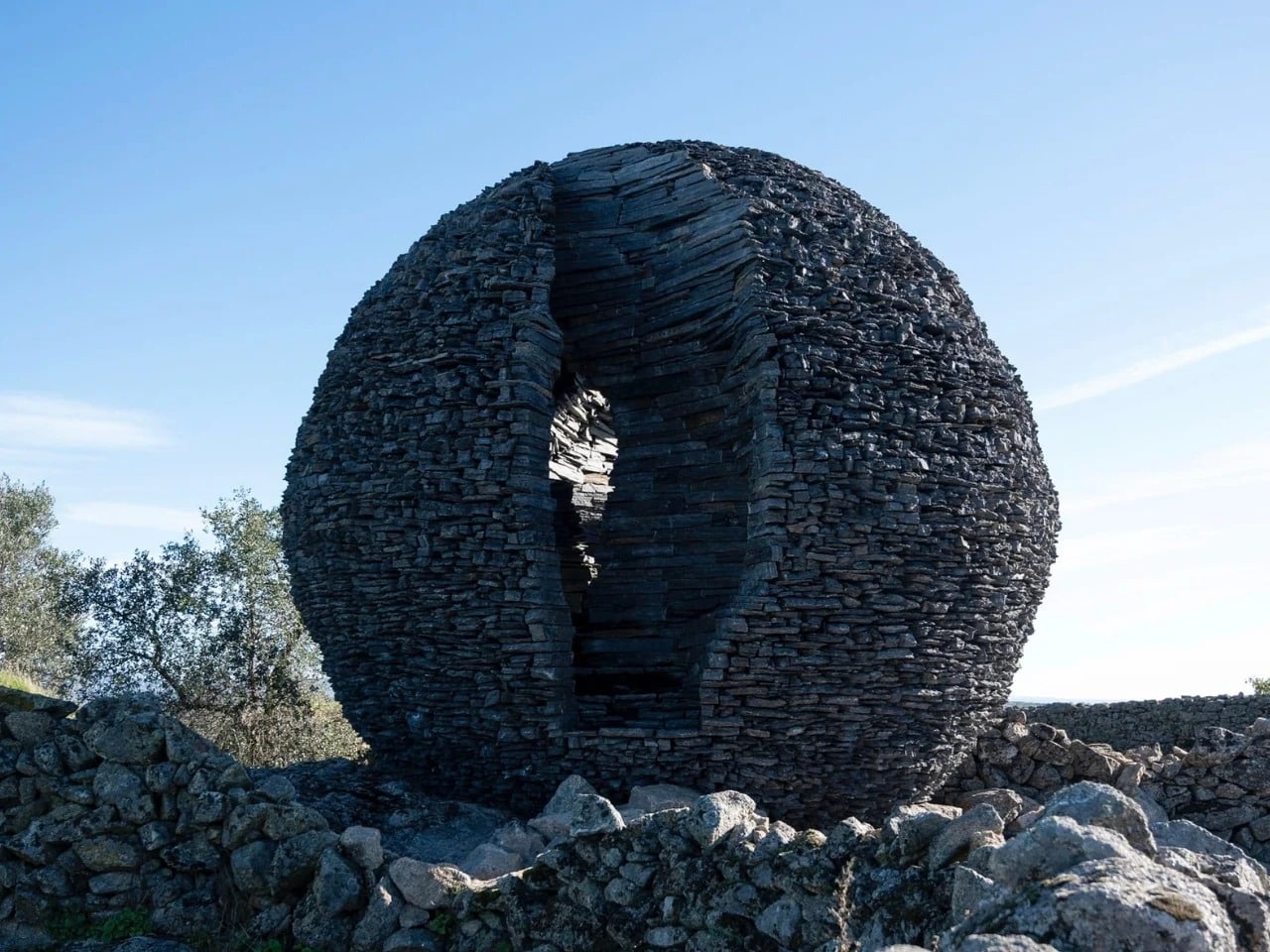 A large spherical structure made of stacked dark schist stone with a narrow vertical opening, set against a clear blue sky.