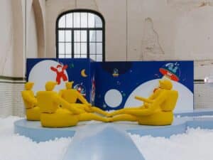 Abstract yellow humanoid sculptures sitting in a circle on a blue platform within a white ball pit at the Little Stranger exhibition.