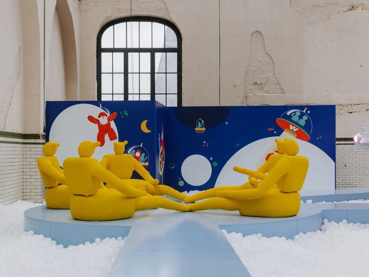 Abstract yellow humanoid sculptures sitting in a circle on a blue platform within a white ball pit at the Little Stranger exhibition.
