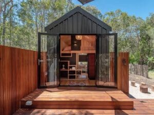 Front view of the Artista tiny house with open black French doors leading to a wooden deck surrounded by trees.