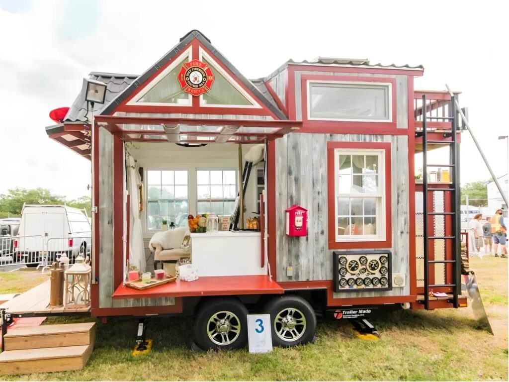Exterior view of a red and grey tiny house designed as a fire station on a trailer with a large open hatch and firefighting emblem.