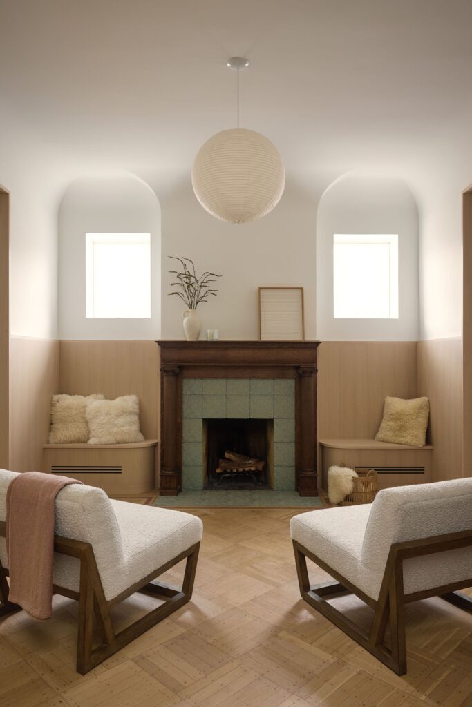 An intimate seating area with two white boucl&eacute; chairs facing a restored wooden fireplace with green tiles.
