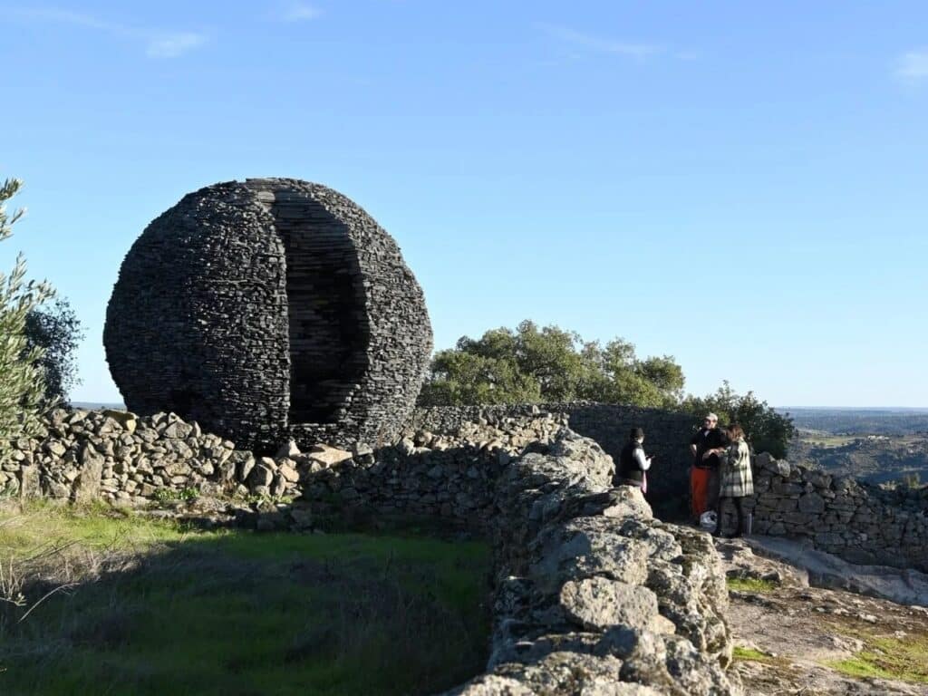 Distant view of the stone sphere in a rocky landscape with visitors walking along a low stone wall under a bright sky.