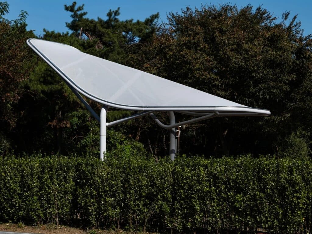Side profile of a single Seoul Wing canopy made of white polyurethane mesh against a backdrop of dense green trees.