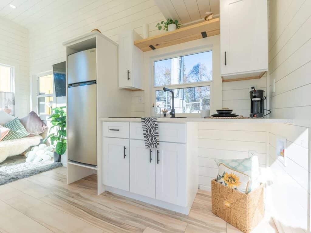 Modern compact kitchen in the Sora 20 tiny home with white cabinetry, stainless steel fridge, and wooden shelving.