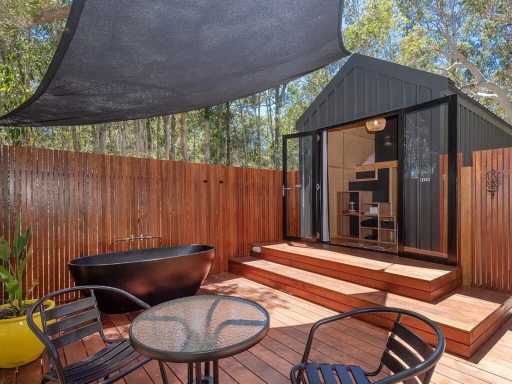 Outdoor lounge area of Artista tiny house featuring a black bathtub, seating set, and a sun shade canopy on a wooden deck.