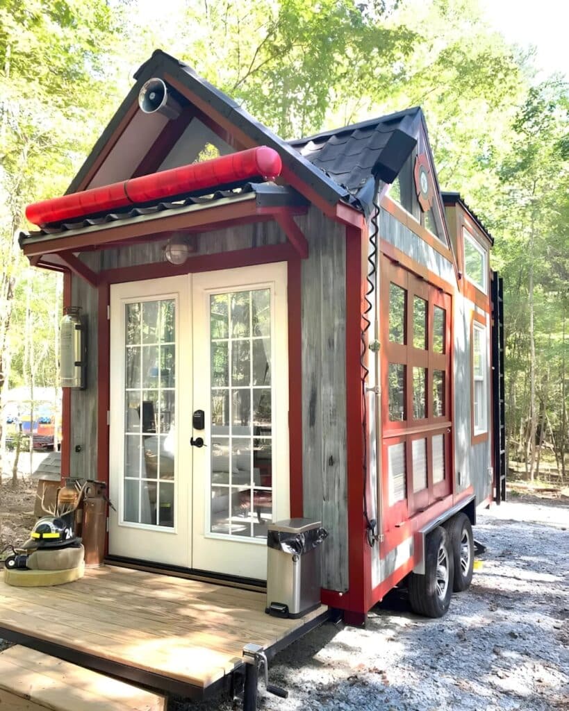 Entrance of the tiny firehouse featuring white French doors, a wooden deck, and a red emergency light bar on the roof.