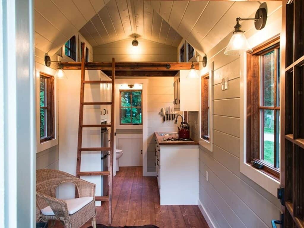 Interior view of the Ynez tiny house looking toward the kitchen and bathroom, featuring a wooden ladder leading to a sleeping loft and pine flooring.
