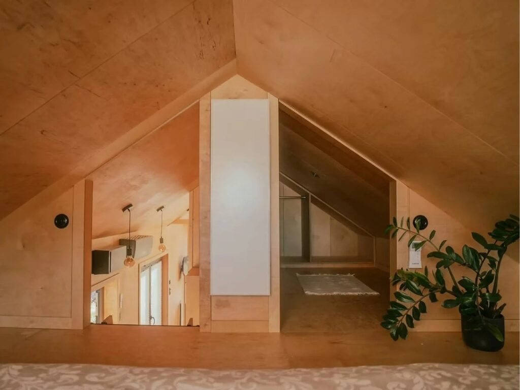 Interior view of a wooden loft space in a tiny house with sloped ceilings and a glimpse into a flexible storage or sleeping area.