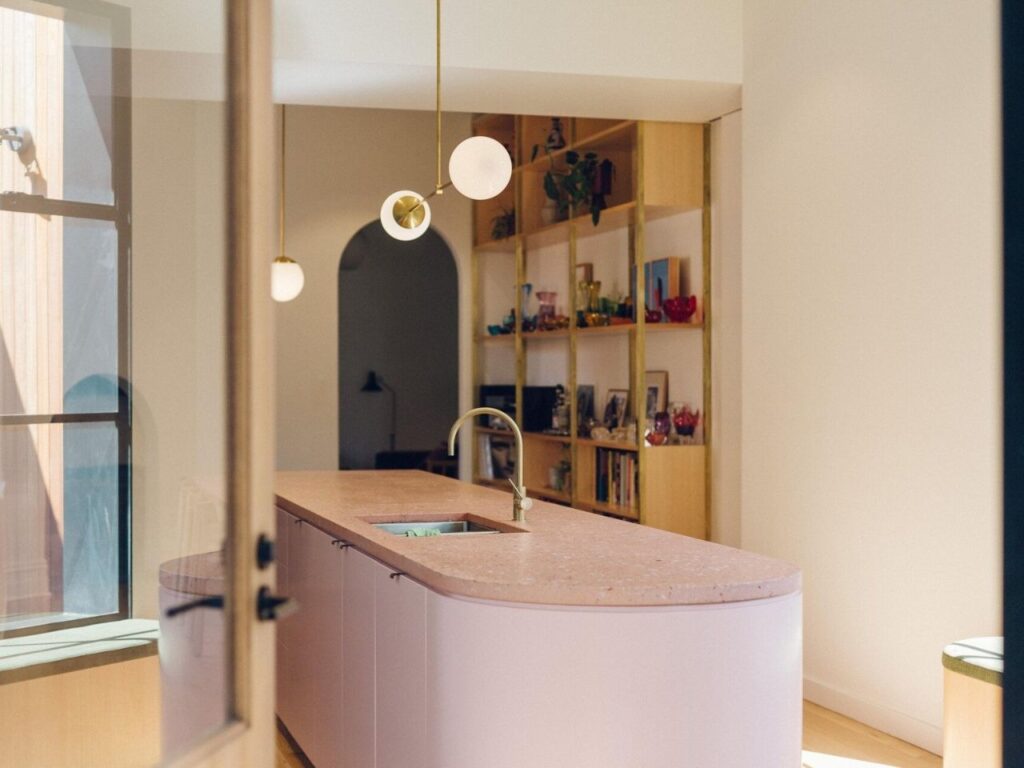 Detail of a curved pink kitchen island with a brass tap, showcasing the connection between the kitchen and the timber shelving in the background.