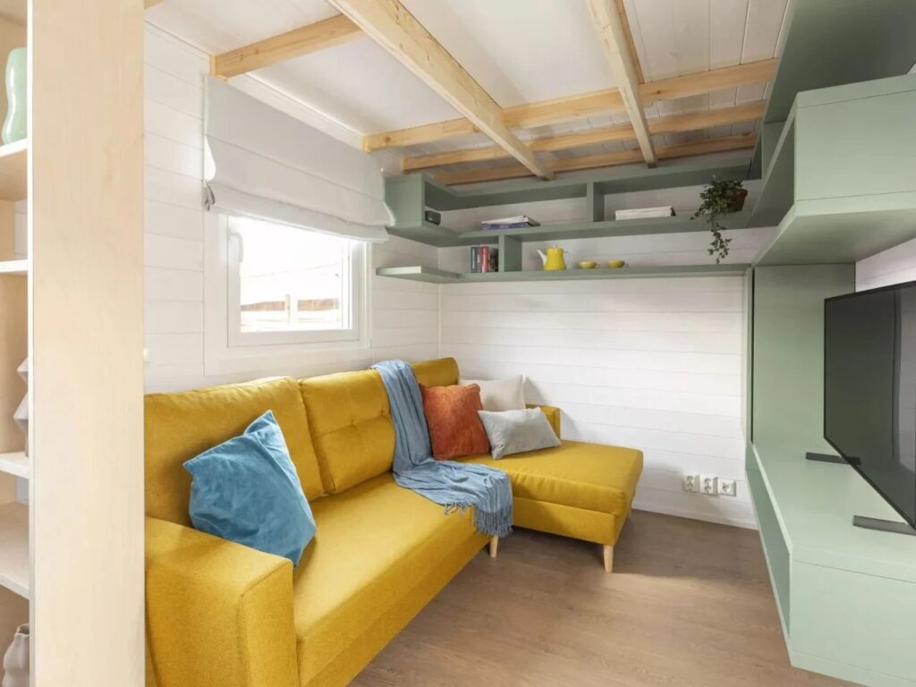 Close-up of a cozy living area in a tiny house with a yellow sectional sofa and built-in sage green wall shelving.