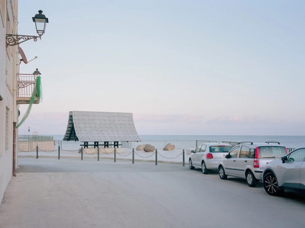 Wide shot of the Asympta pavilion located on a public waterfront plaza with parked cars and traditional buildings in the foreground.
