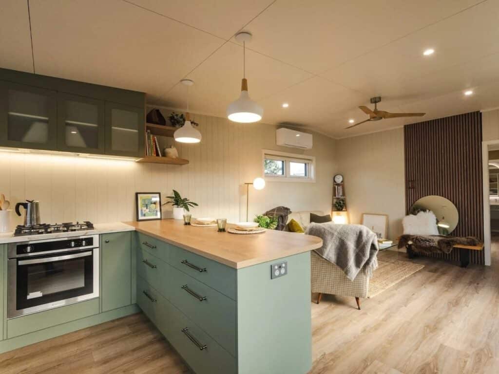 Wide shot of a functional tiny house kitchen with sage green cabinetry and a large wooden island serving as a dining area.
