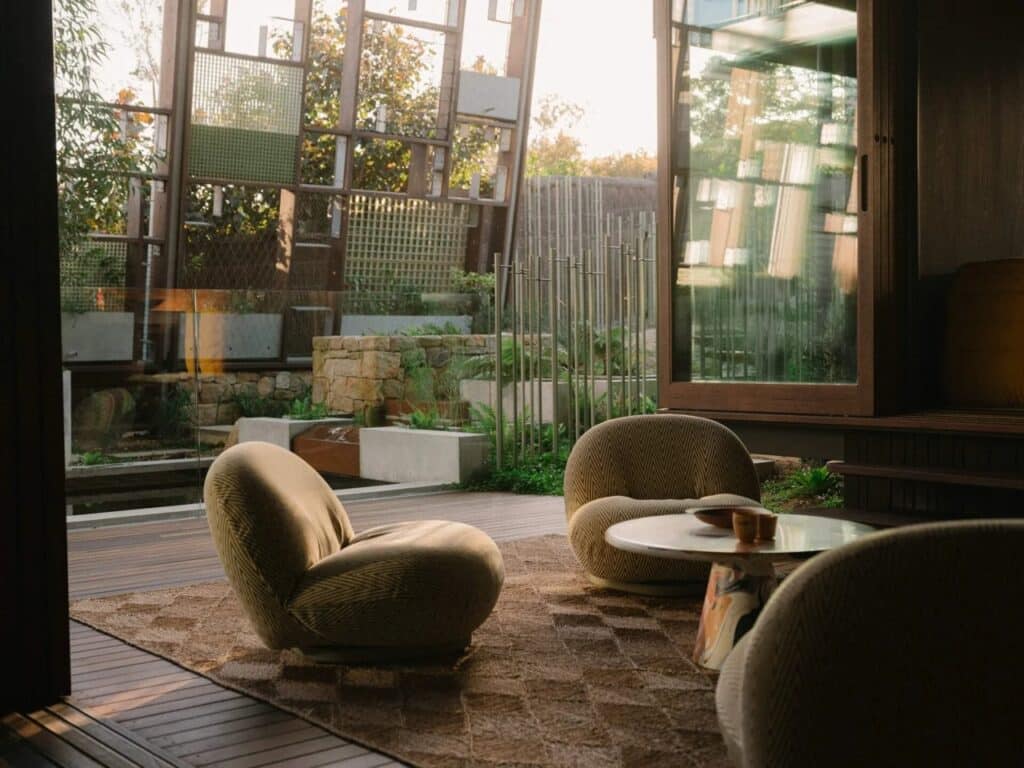 Sun-drenched interior lounge area of Holocene House featuring organic-shaped seating, a textured rug, and large glass doors opening to a garden.