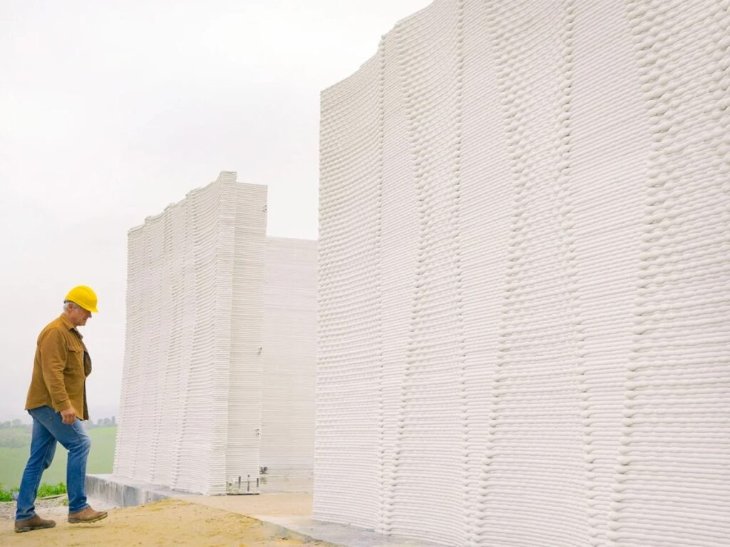 An architect walking past the textured, layered 3D-printed walls made of a lime-based mixture.