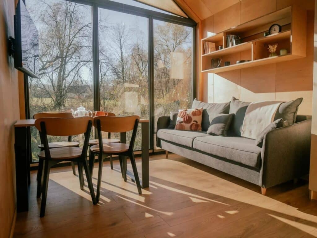 Sunlit living area in Zenith tiny house with a grey sofa, wooden dining table, and massive windows overlooking a forest.