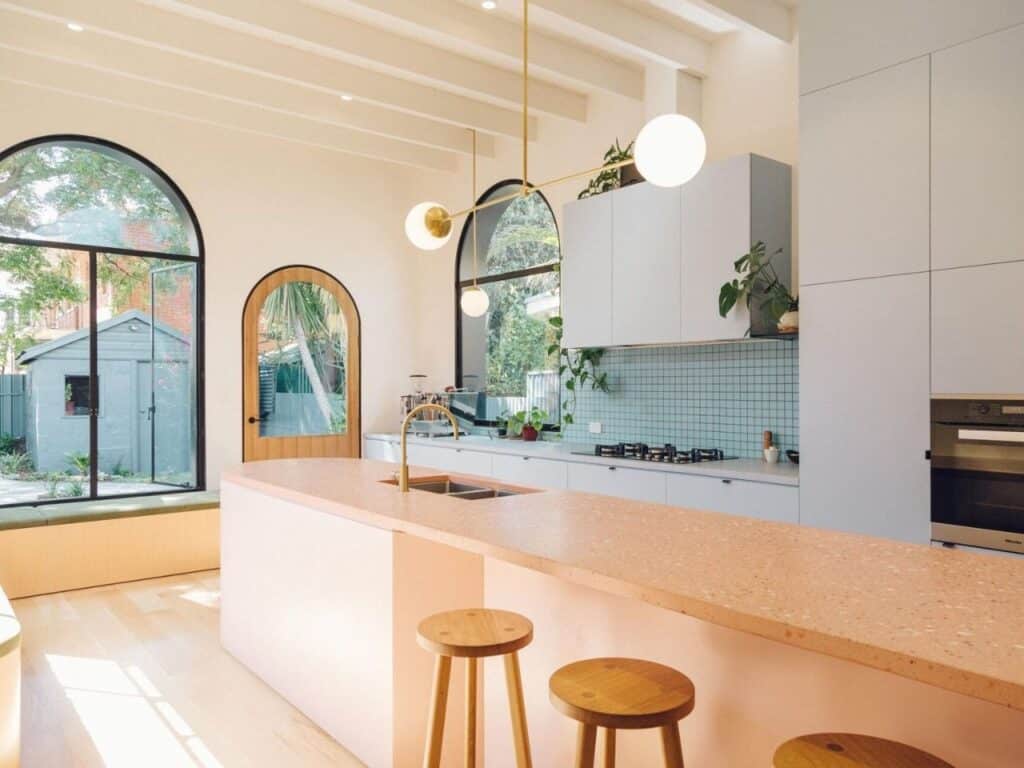 Modern kitchen in the Adelaide cottage with light blue tiled backsplash, white cabinets, and a long pink island with wooden stools.